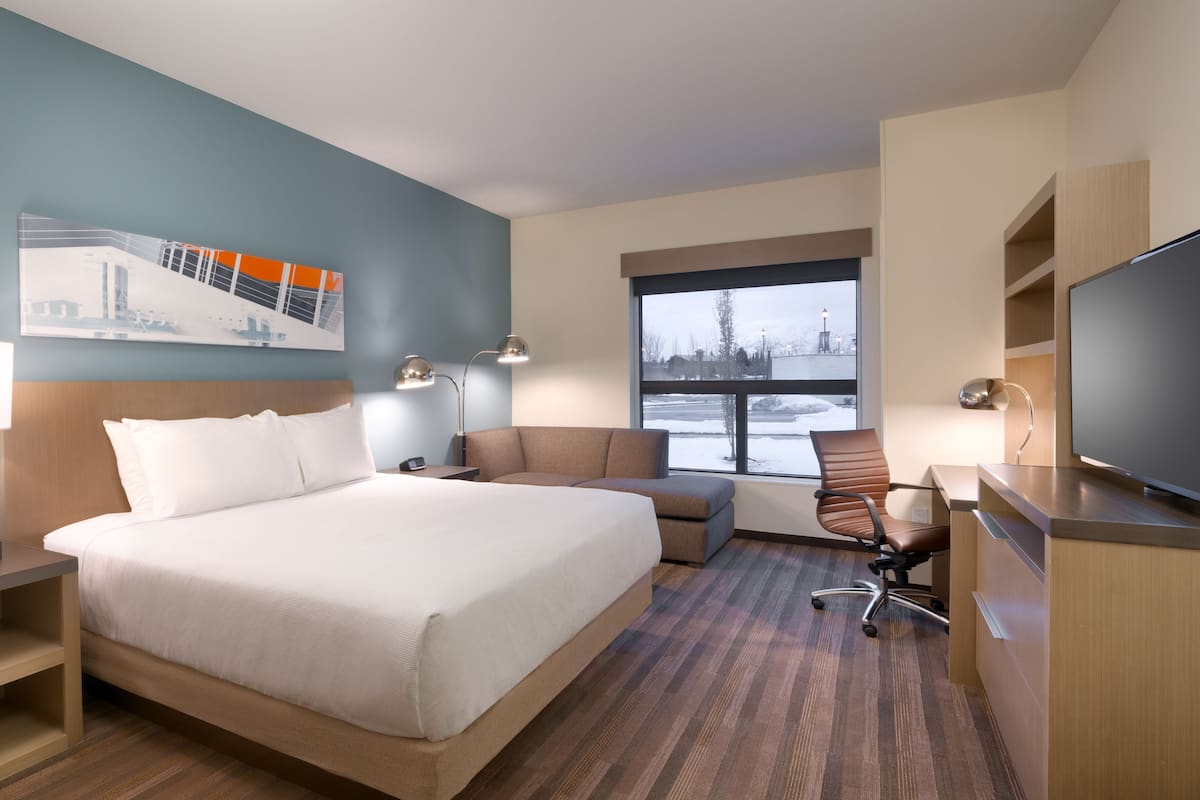 Ein Hotelzimmer mit einem großen Bett, einem Schreibtisch mit Flachbildfernseher, einem Sessel, einer Schreibtischlampe und einem Fenster mit Blick auf eine verschneite Landschaft.