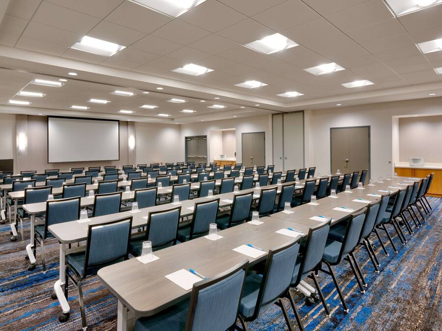 A conference room with rows of tables and chairs, a projector screen, and a kitchenette area.