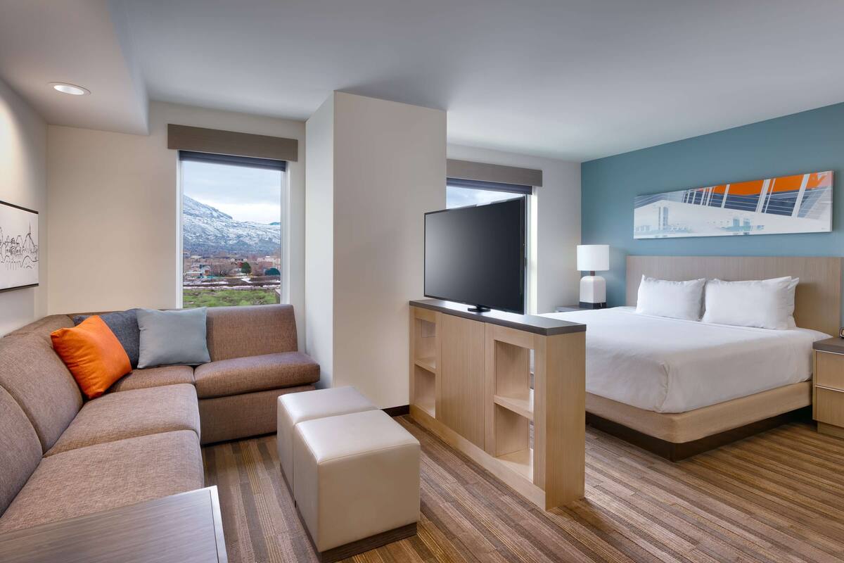 Ein modernes Hotelzimmer mit Bett, Sofa, Fernseher und Blick auf Berge.