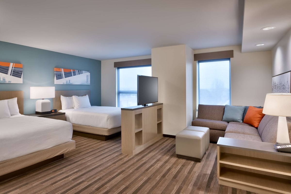 Ein Hotelzimmer mit zwei Betten, einem Fernseher, einem Sofa und einem Schreibtisch mit einer Lampe.