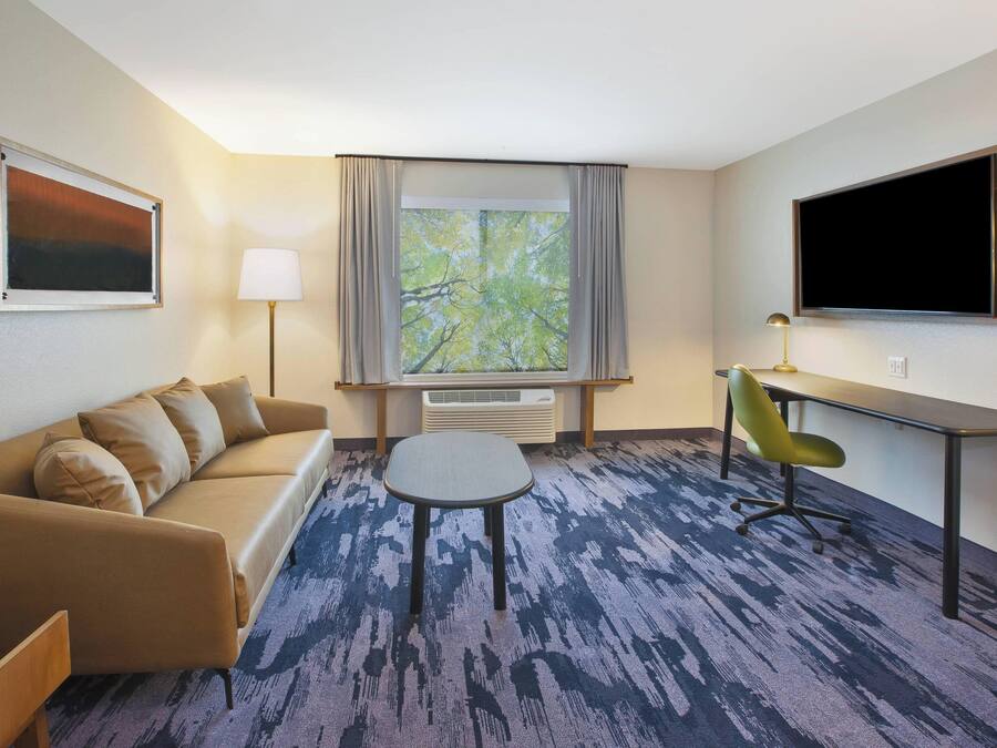 A hotel room with a sofa, a small table, a desk with a chair, a TV, and a window with curtains.
