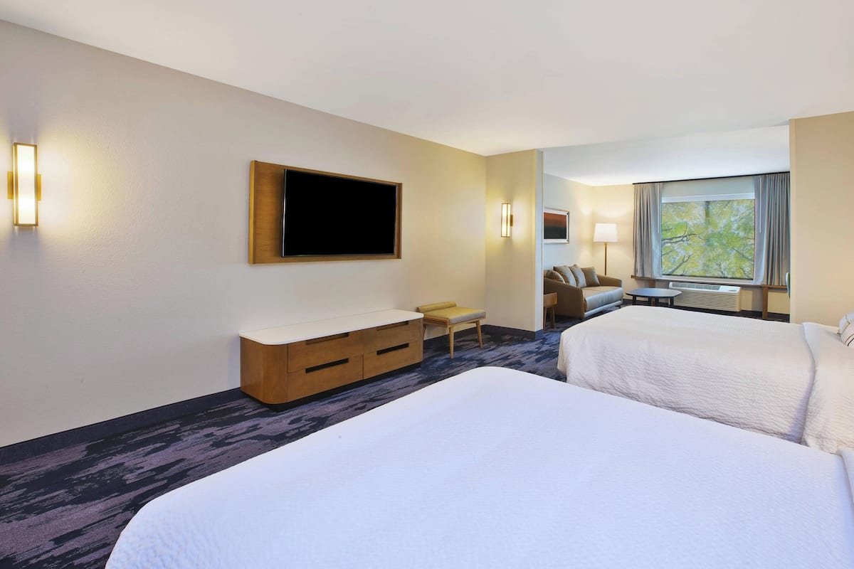 A hotel room with two beds, a TV mounted on the wall, a small seating area with a chair and table, and a window with a view of trees.