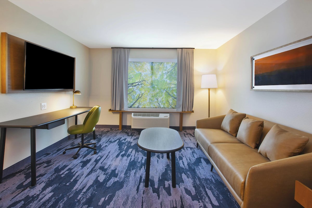 A hotel room with a desk, chair, sofa, TV, window with curtains, and a lamp.