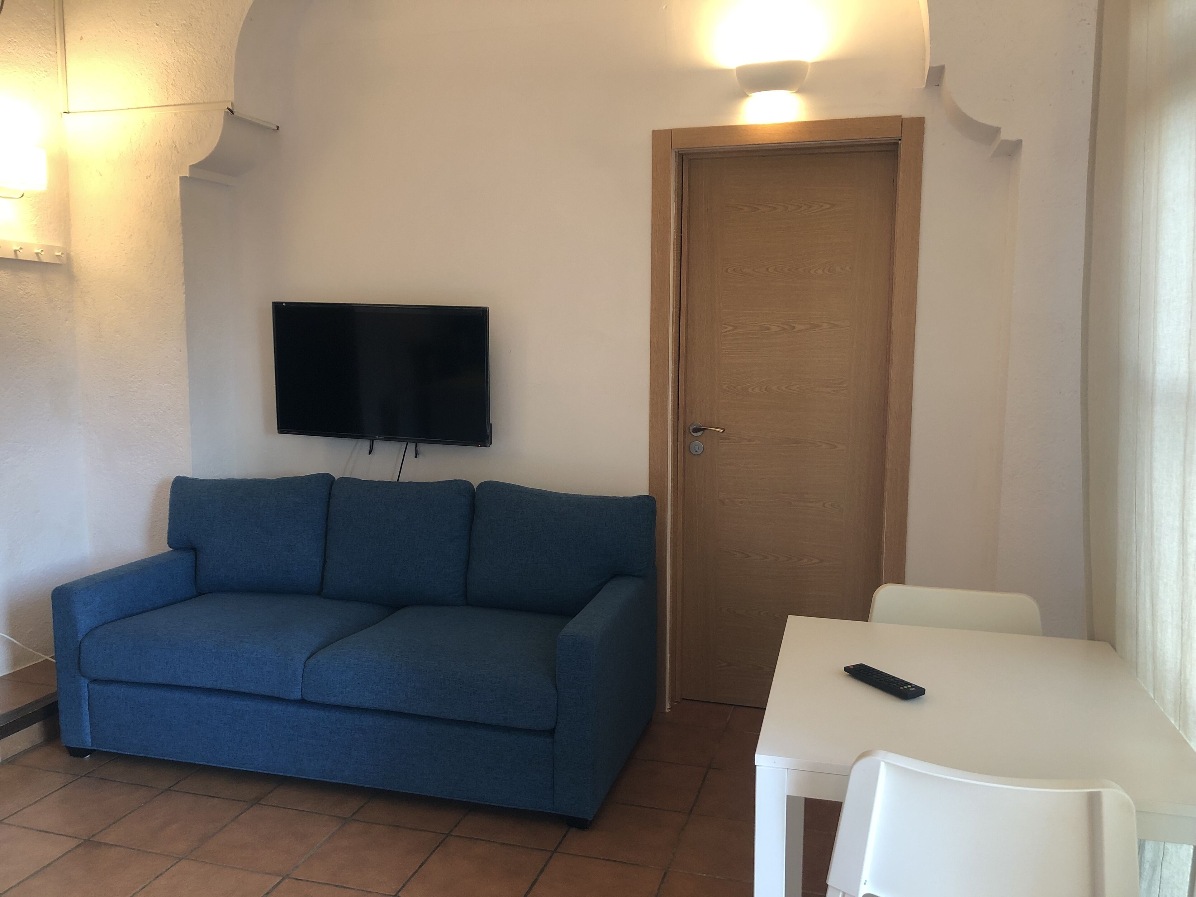 Una habitación con un sofá azul, una mesa blanca, un televisor montado en la pared y una puerta de madera.