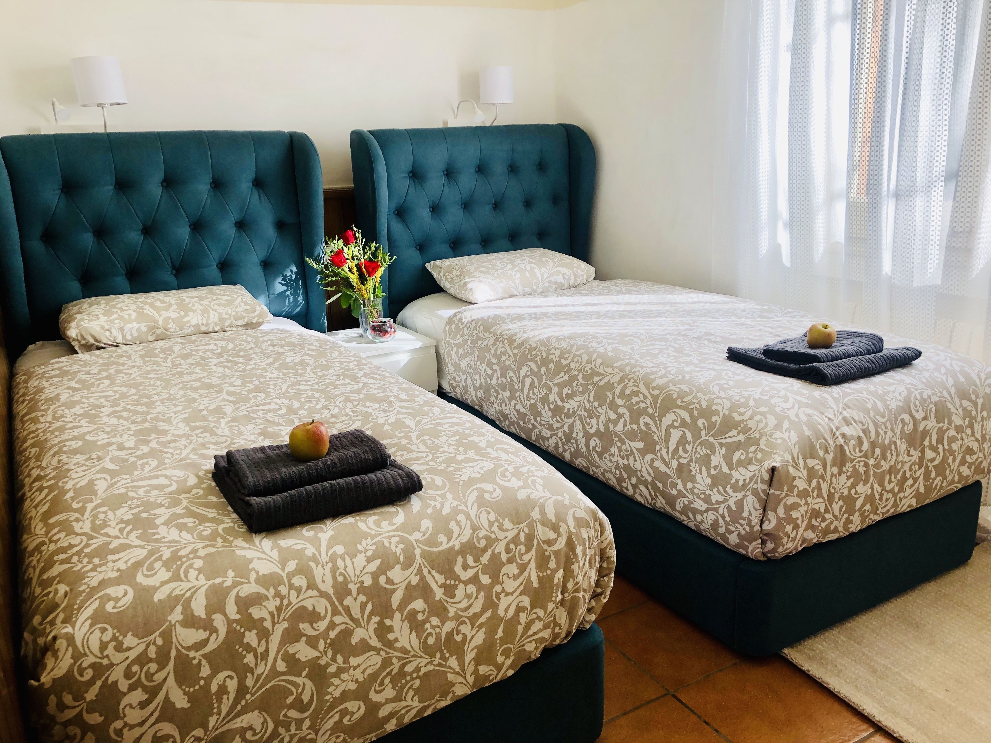 A hotel room with two beds, each with a tufted headboard, a bedside table with a vase of flowers, and a window with sheer curtains.