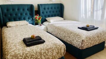 A hotel room with two beds, each with a tufted headboard, a bedside table with a vase of flowers, and a window with sheer curtains.