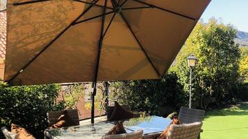 Une terrasse extérieure avec une table en verre et des chaises en osier, protégée du soleil par un grand parasol.