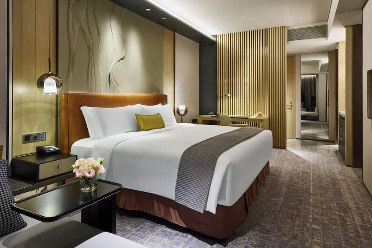 A hotel room with a large bed, bedside tables, a desk, a chair, a desk lamp, a vase with flowers, and a mirror.