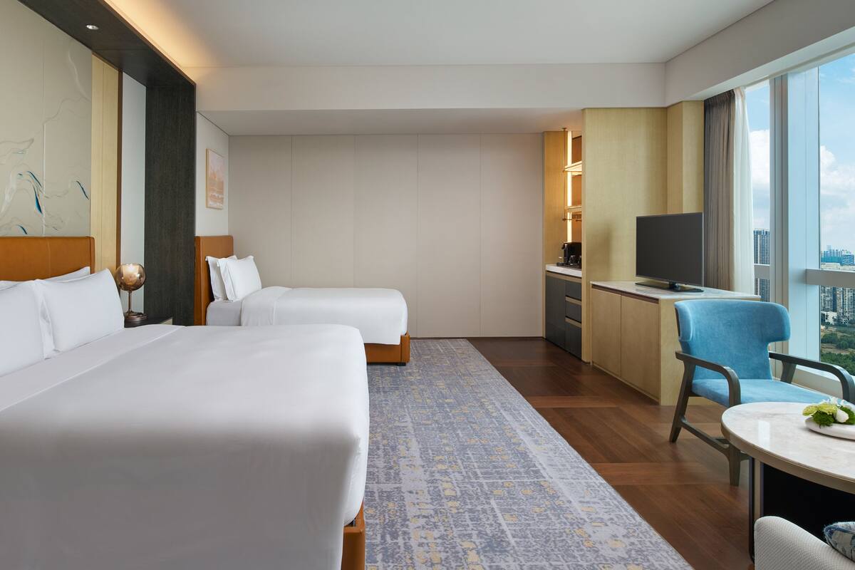 A hotel room with two beds, a desk, a TV, and a view of the city.