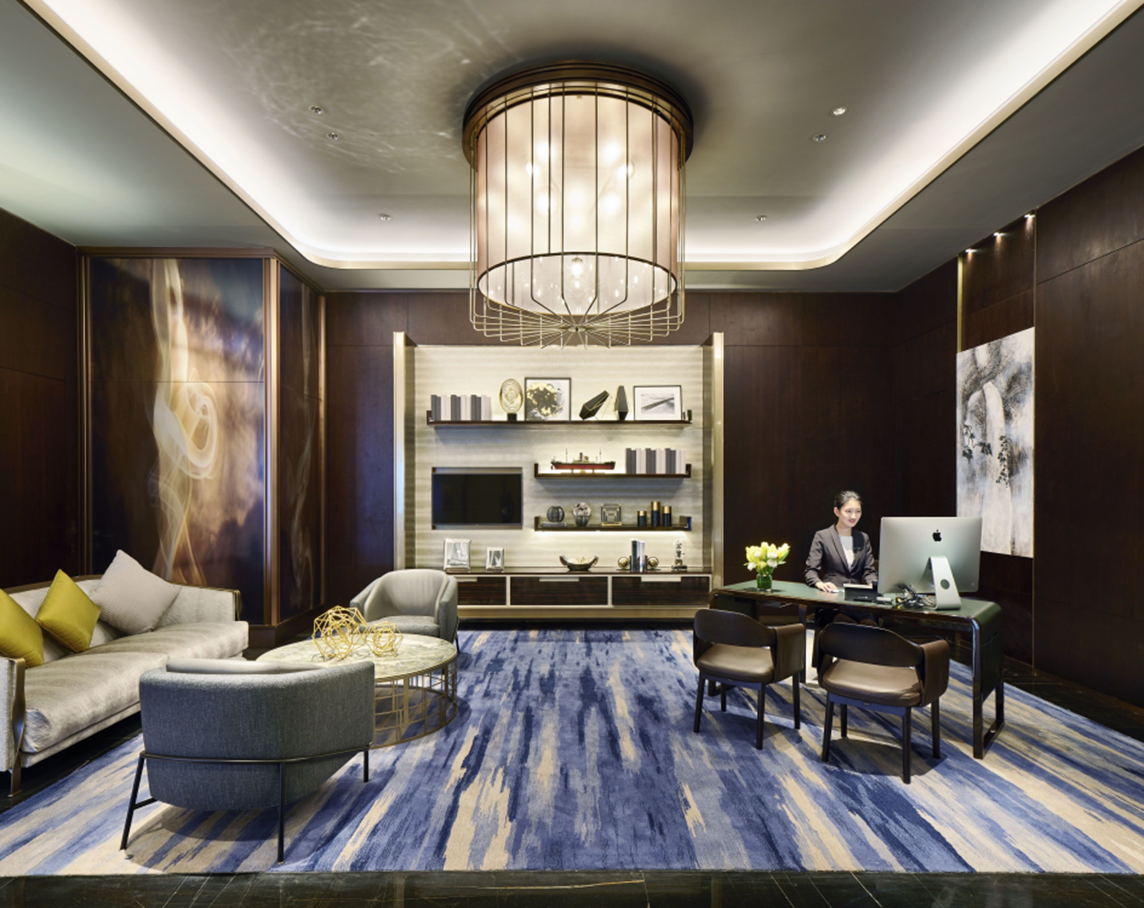 A modern hotel lobby with a large chandelier, a sofa with cushions, a coffee table, a desk with a computer, and a bookshelf.