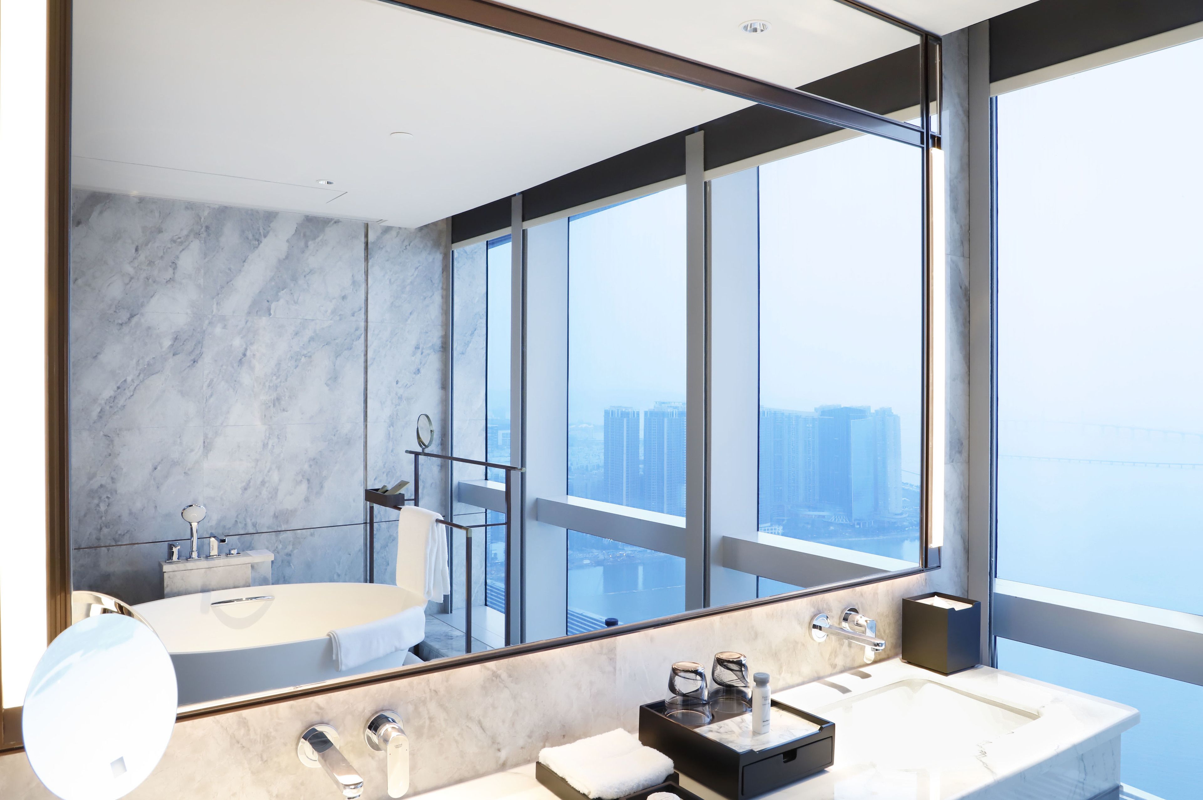 Un baño moderno con un espejo grande, dos lavamanos y una bañera. Ofrece vistas de la ciudad a través de amplias ventanas.