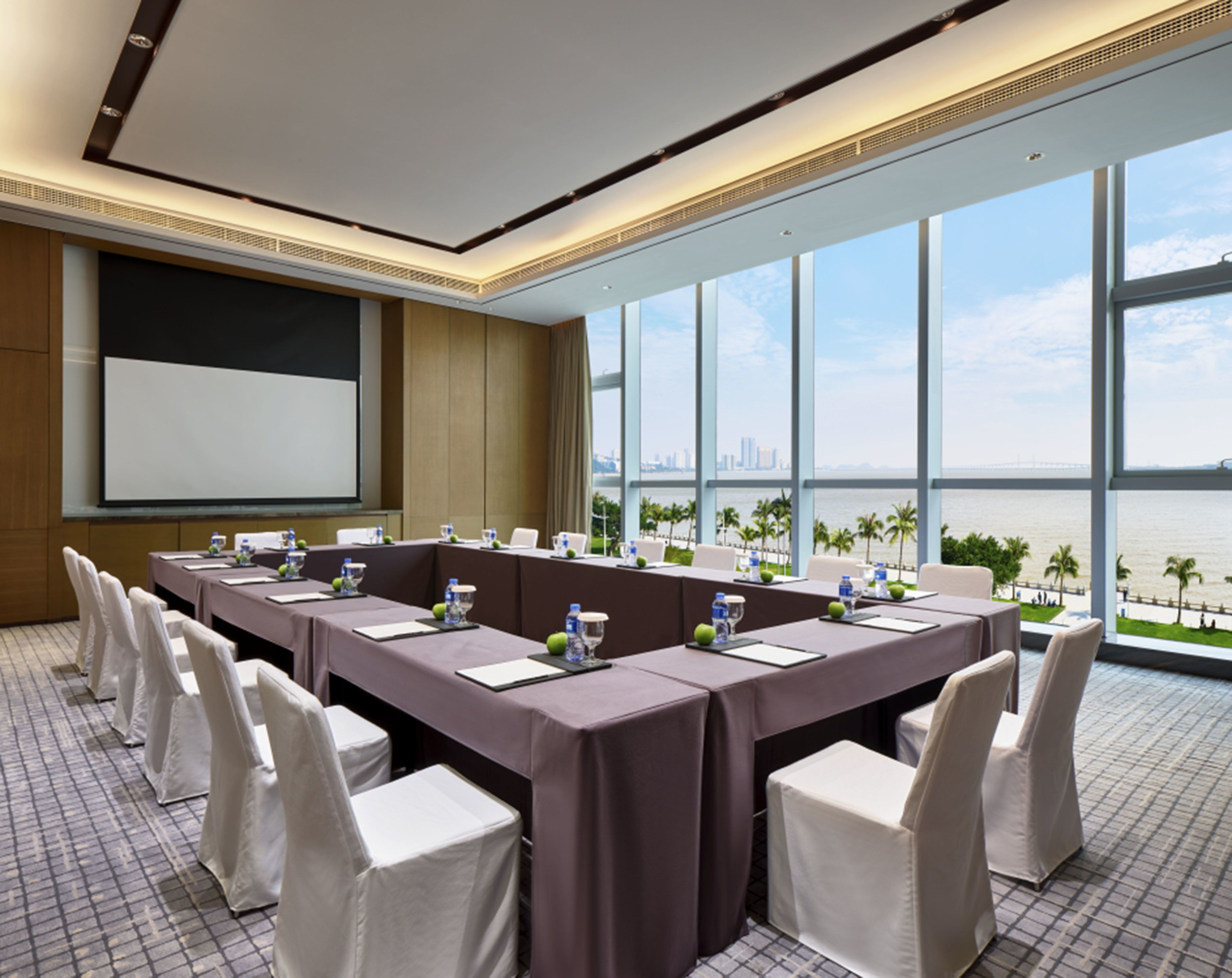 Un salón de conferencias con una larga mesa, sillas blancas, una pantalla de proyector y vistas al agua y al horizonte urbano.