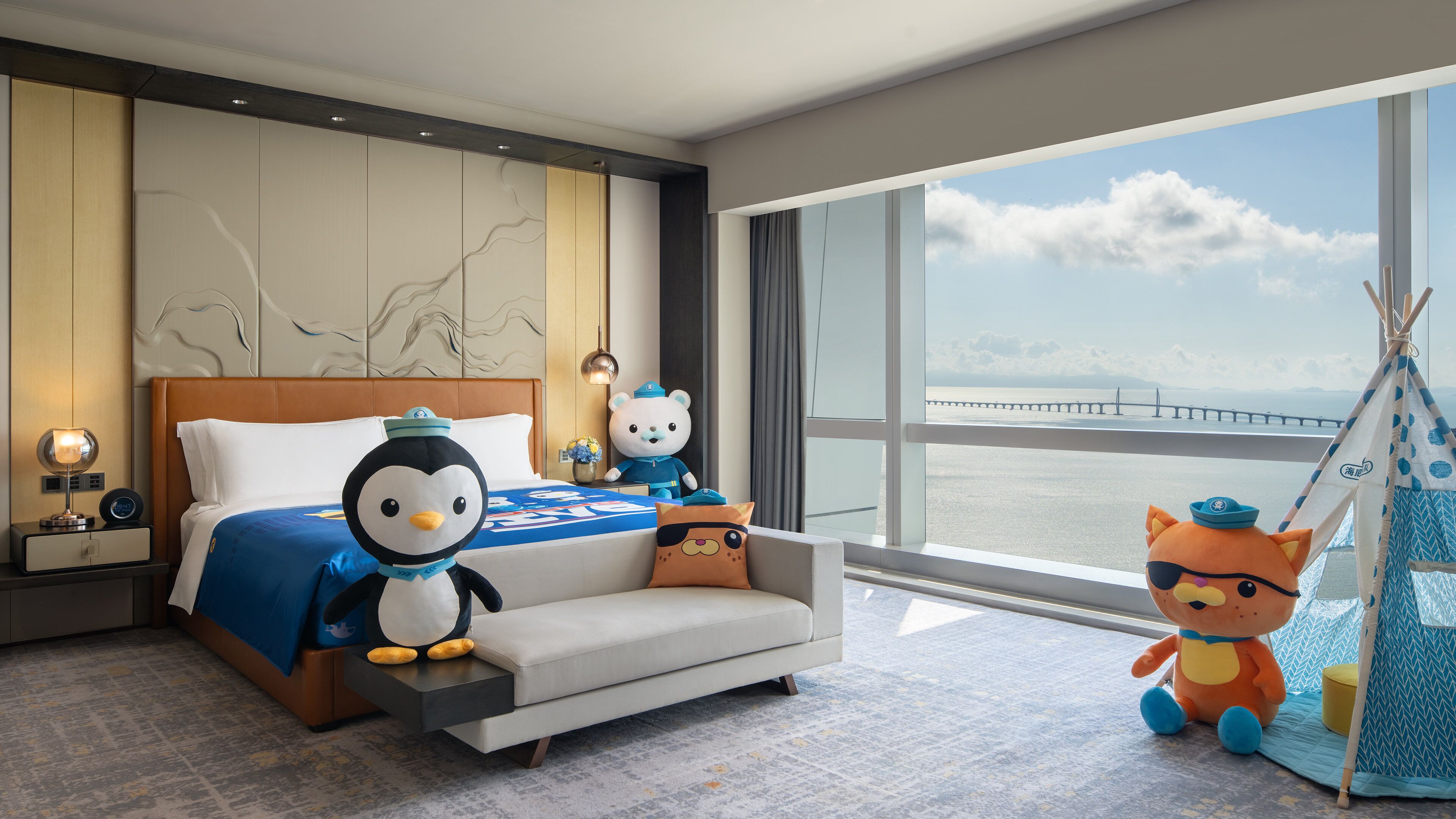Habitación de hotel con una cama grande, un sofá, una mesita de noche con lámpara, un oso de peluche, un peluche de pingüino, otro oso de peluche y un tipi con un peluche.