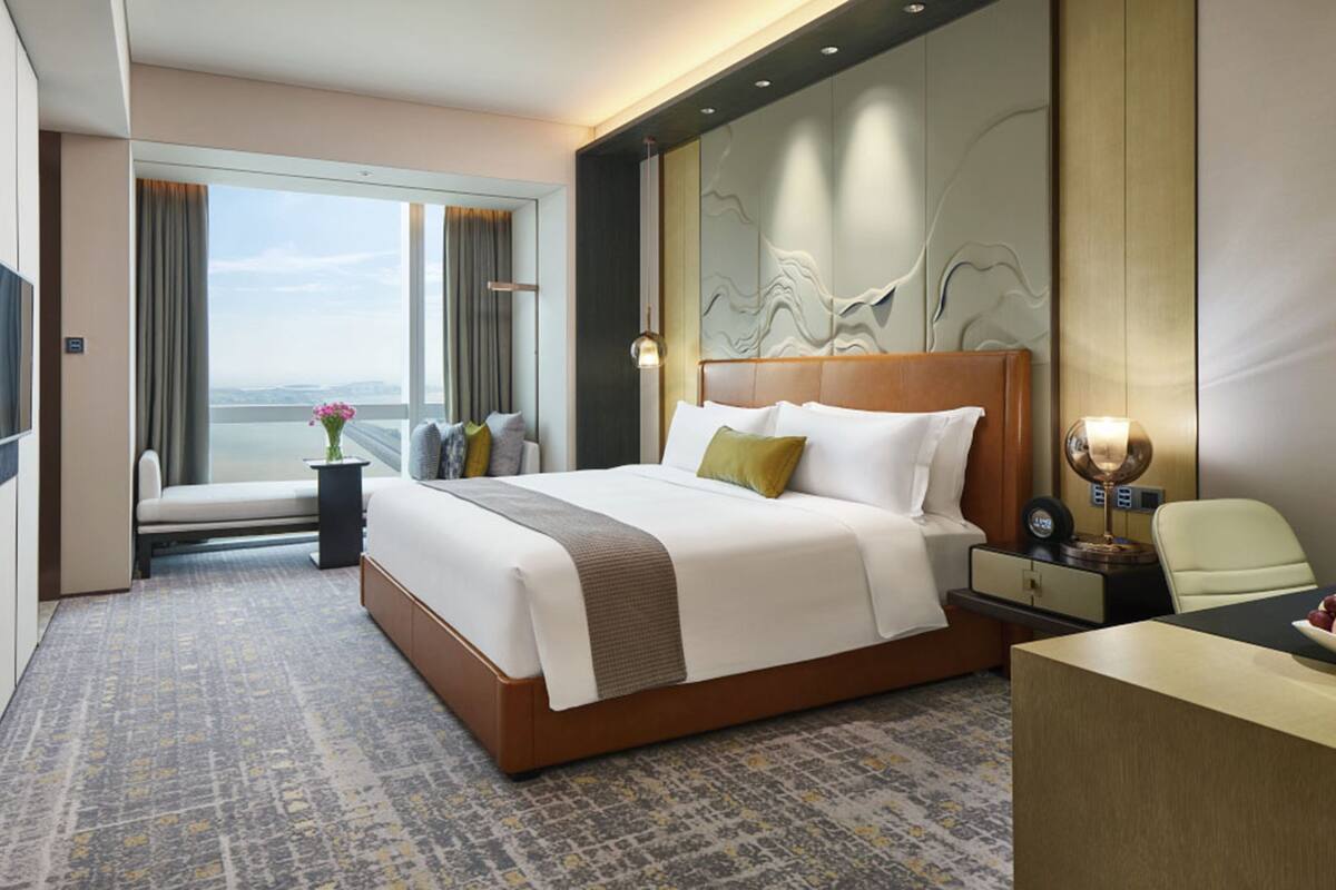 A hotel room with a large bed, a desk, a chair, a nightstand, a lamp, and a view of the city.