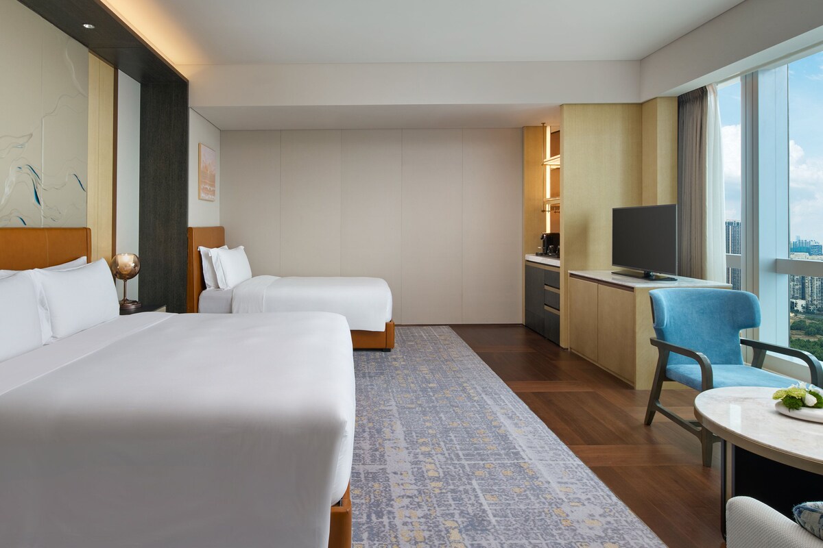 Habitación de hotel con dos camas, un escritorio, una silla, un televisor y un ventanal con vistas a la ciudad.