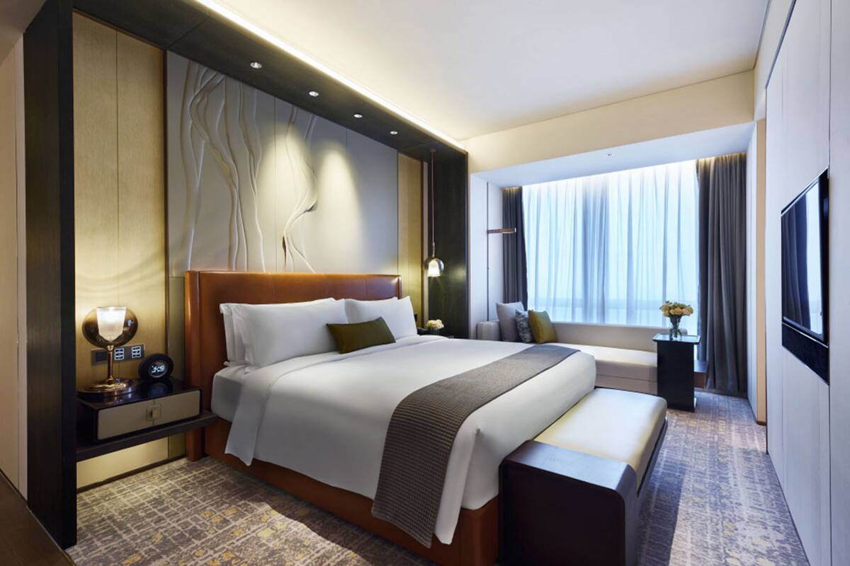 A modern hotel room with a large bed, a bedside table with a lamp, a bench, a TV, and a large window with curtains.