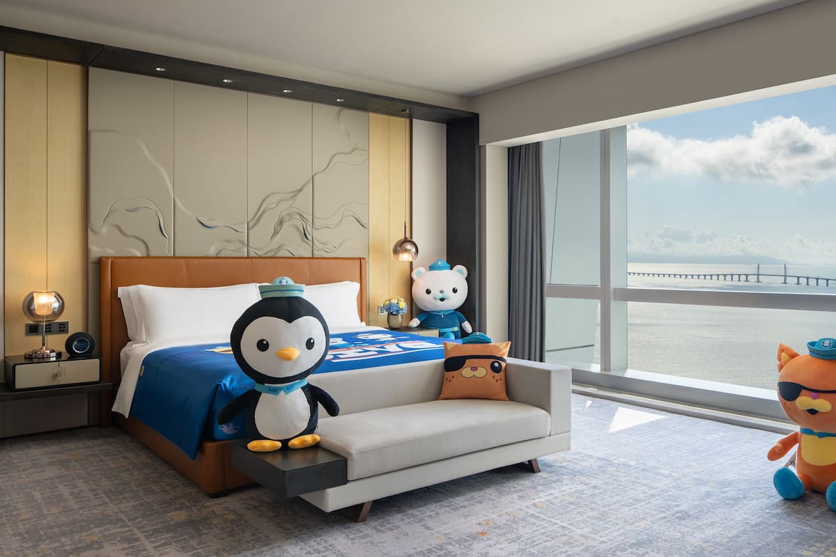 A hotel room with a large bed, a sofa, a nightstand with a lamp, a teddy bear, a penguin plush toy, a bear plush toy, and a teepee with a stuffed animal.