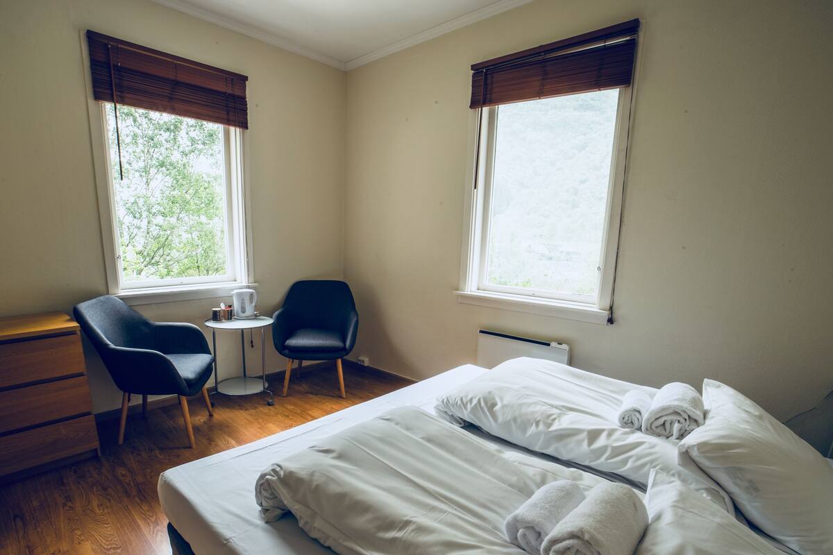 Ein Hotelzimmer mit einem Bett, zwei Sesseln, einem kleinen Tisch und zwei Fenstern mit Jalousien.