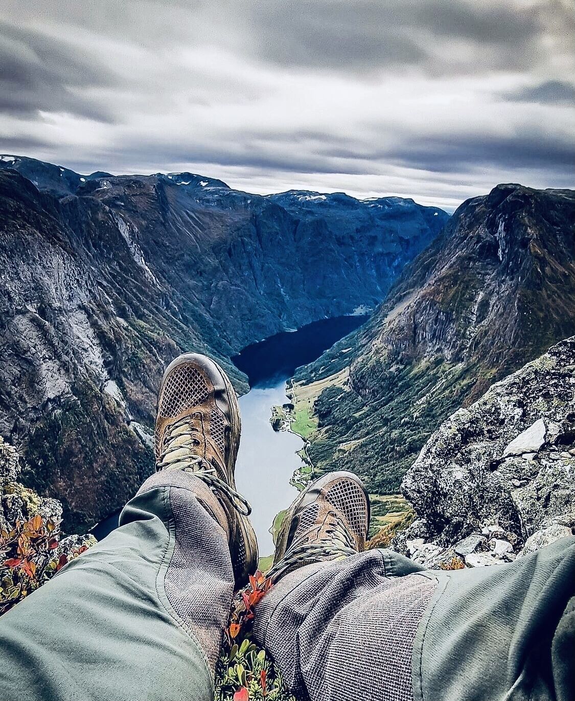 Una persona sentada con las piernas cruzadas, contemplando un valle con un lago y montañas al fondo.