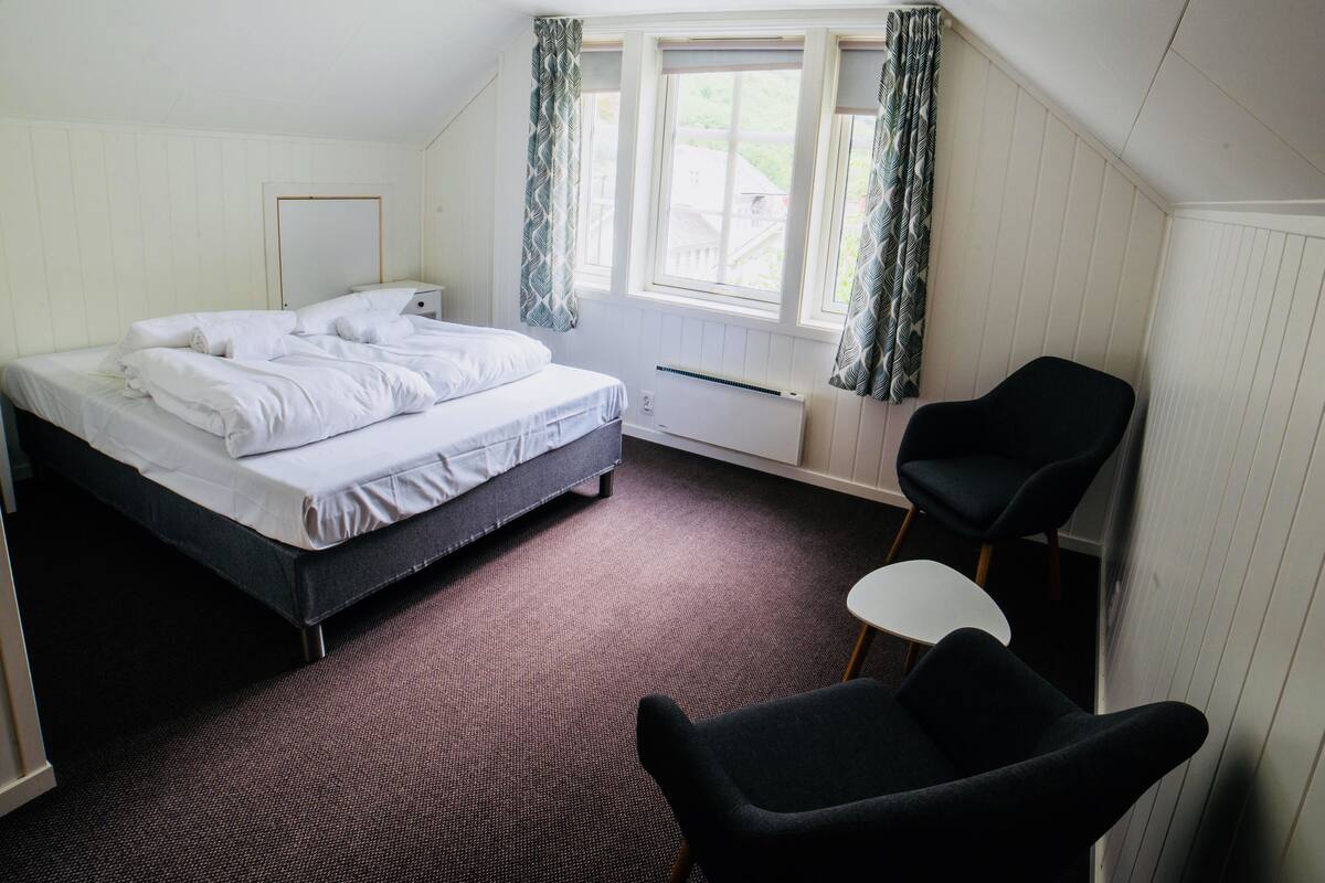 Ein Hotelzimmer mit Bett, Stuhl, kleinem Tisch und einem Fenster mit Vorhängen.