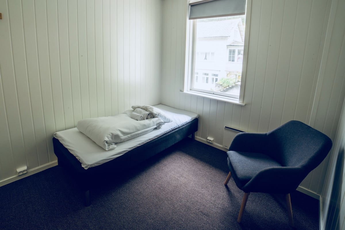 Ein kleines Zimmer mit einem Einzelbett, einem Fenster, einem Sessel und Teppichboden.