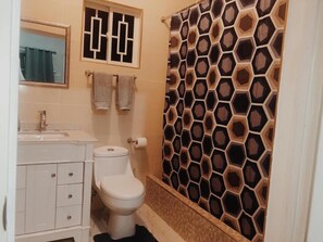 Un baño con inodoro, lavamanos y espejo. Se observa una cortina de ducha con diseño estampado y una ventana con diseño geométrico.
