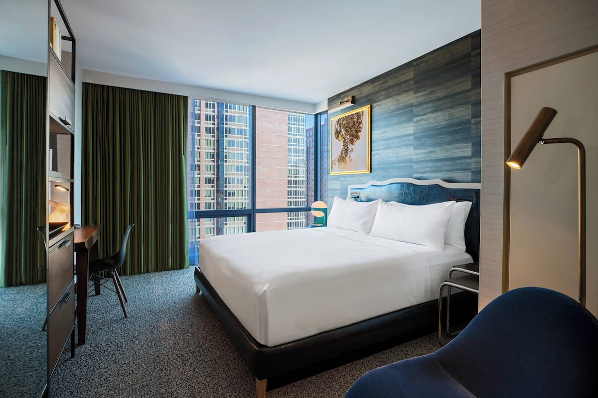 A hotel room with a large bed, a desk, a chair, a nightstand, and a view of buildings through large windows.