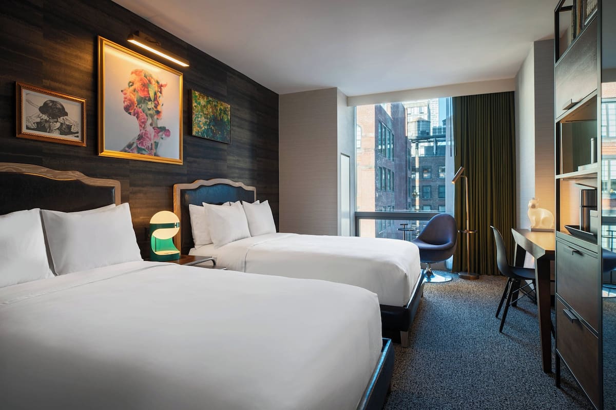 A hotel room with two beds, a desk, a chair, a lamp, and a large window with a view of buildings.