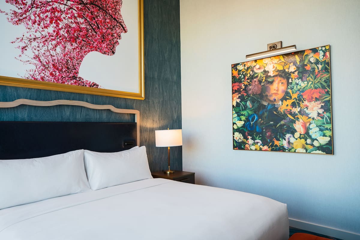 A hotel room with a bed, a nightstand with a lamp, and two large floral paintings on the wall.