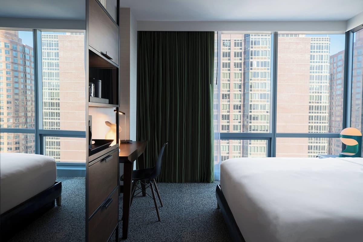 A hotel room with two beds, a desk, and a chair. There is a large window with a view of tall buildings.