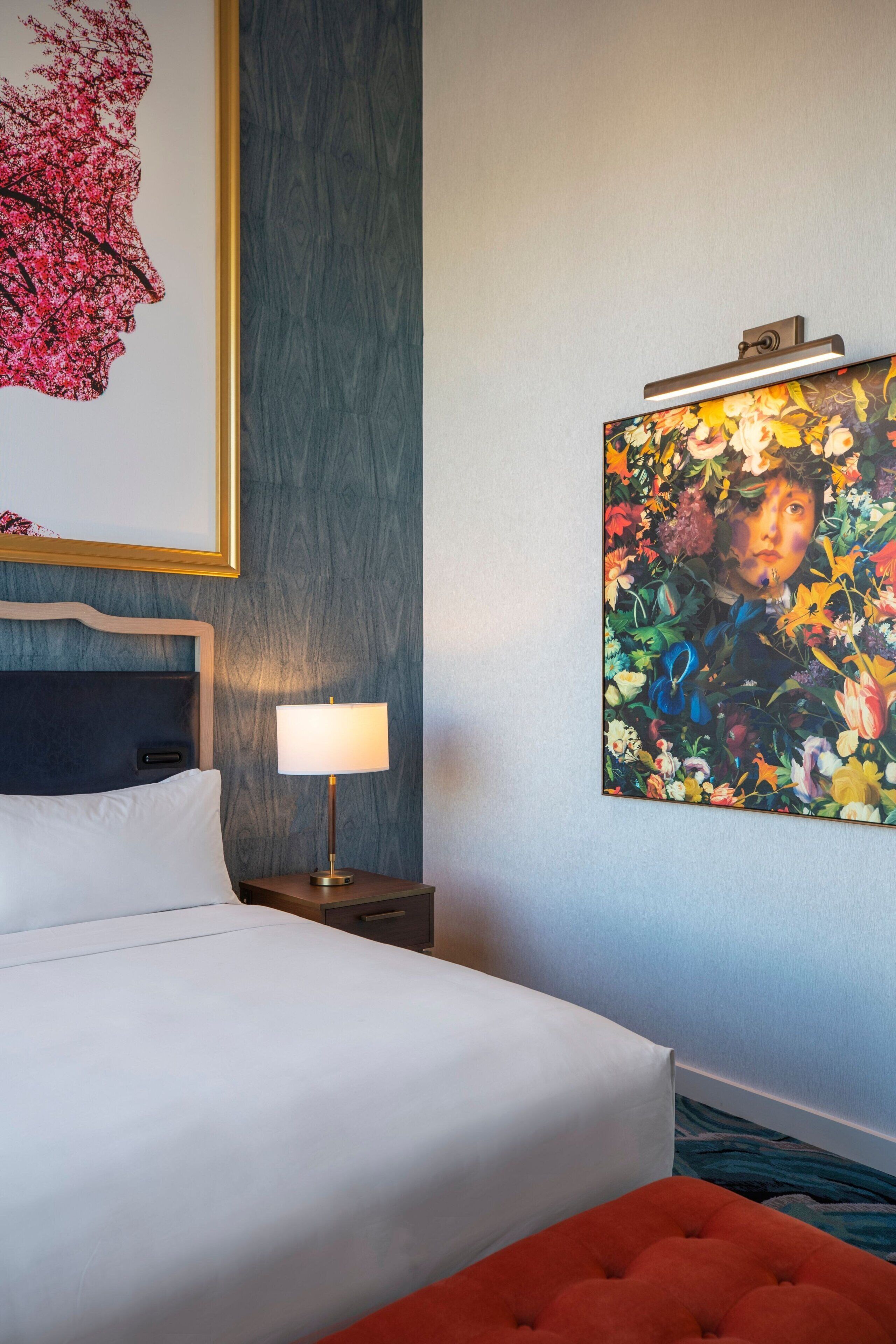 A hotel room with a bed, a nightstand with a lamp, and two wall-mounted artworks.