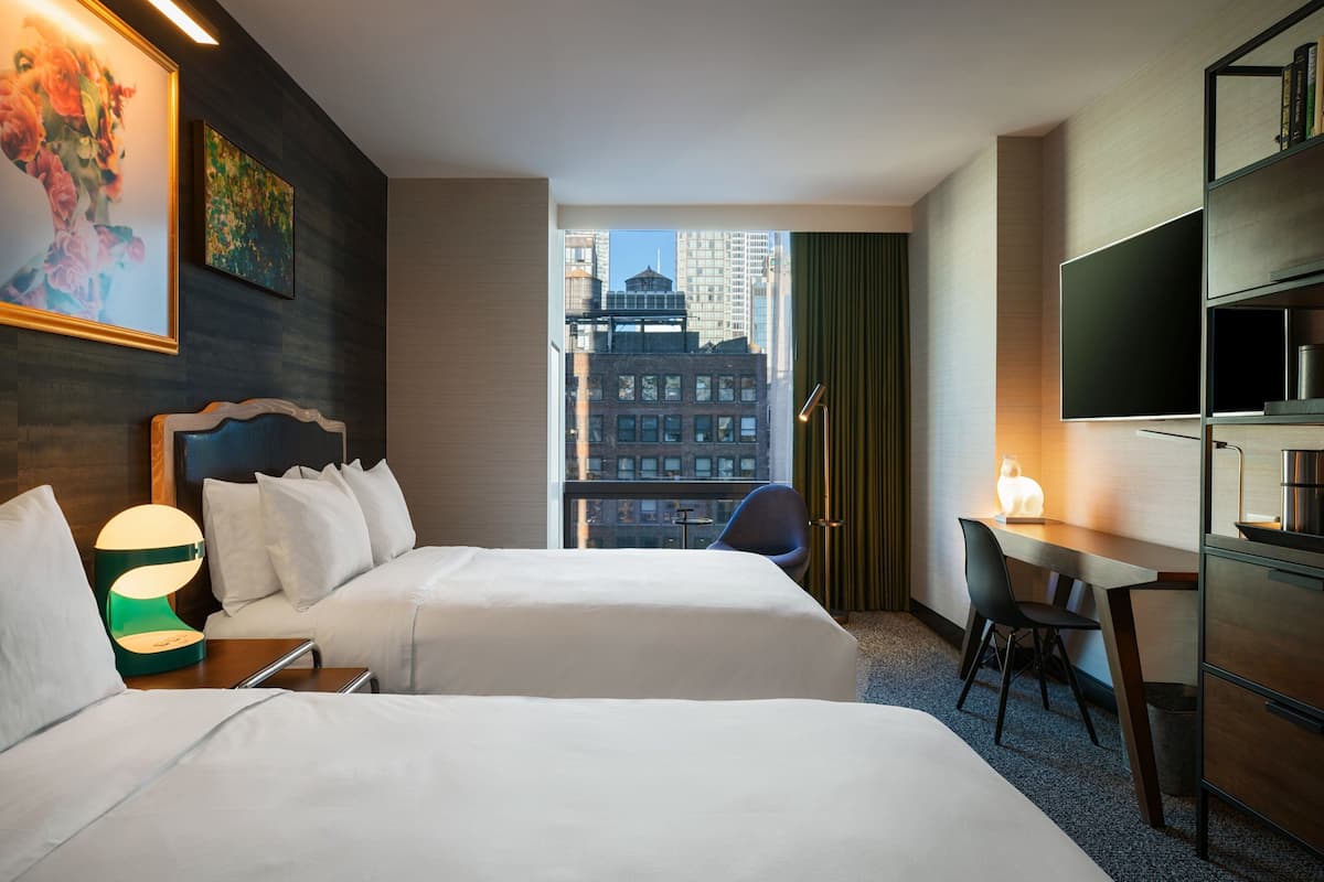 A hotel room with two beds, a desk, and a chair. There is a large window with a view of buildings outside.