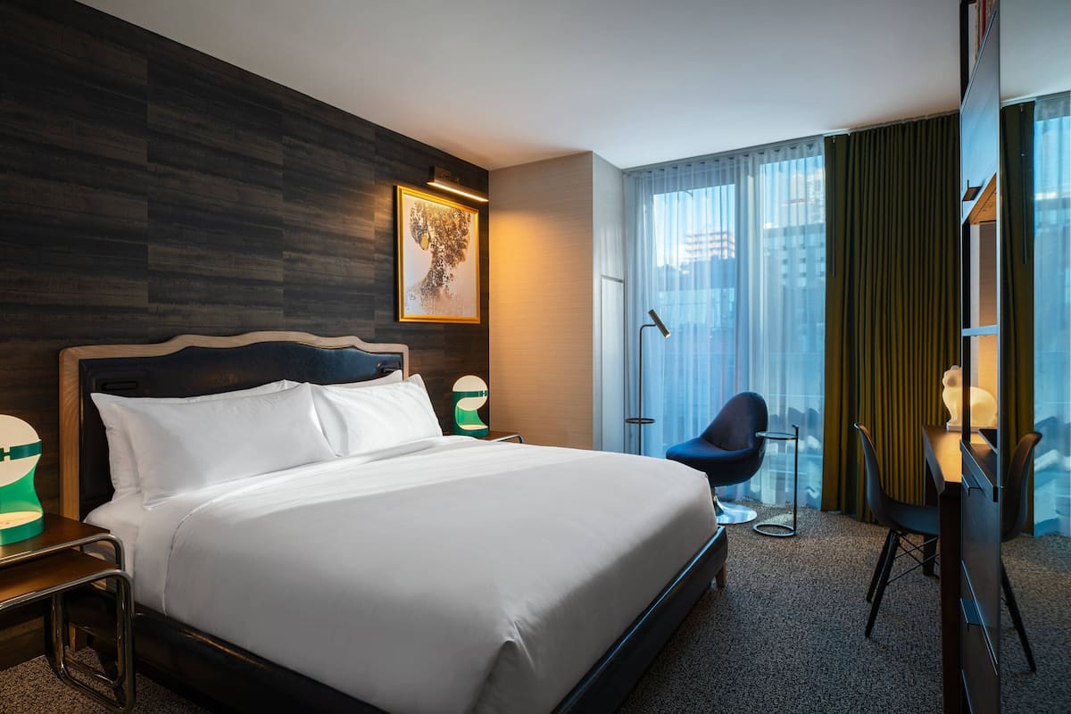 A hotel room with a large bed, a desk with a chair, a desk lamp, a framed picture on the wall, and a view of the city through the window.