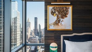A hotel room with a large window, a framed artwork, a bedside table with a lamp, and a neatly made bed.