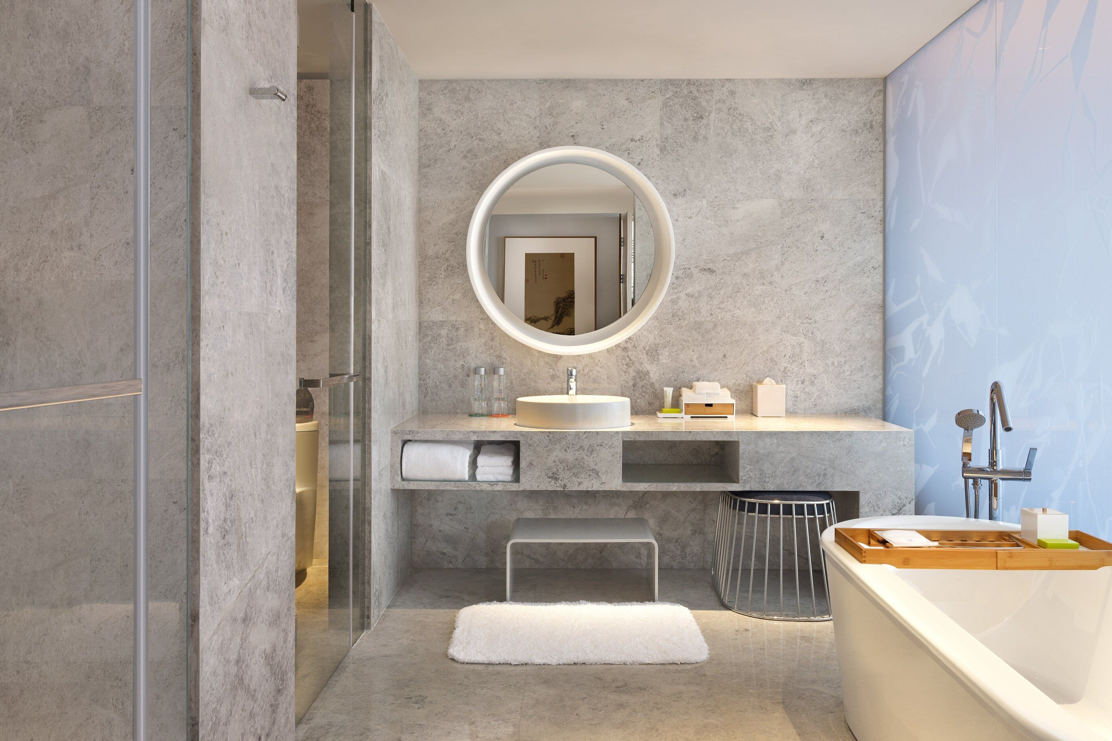 A modern bathroom with a marble sink, a round mirror, a bathtub, and a bench.