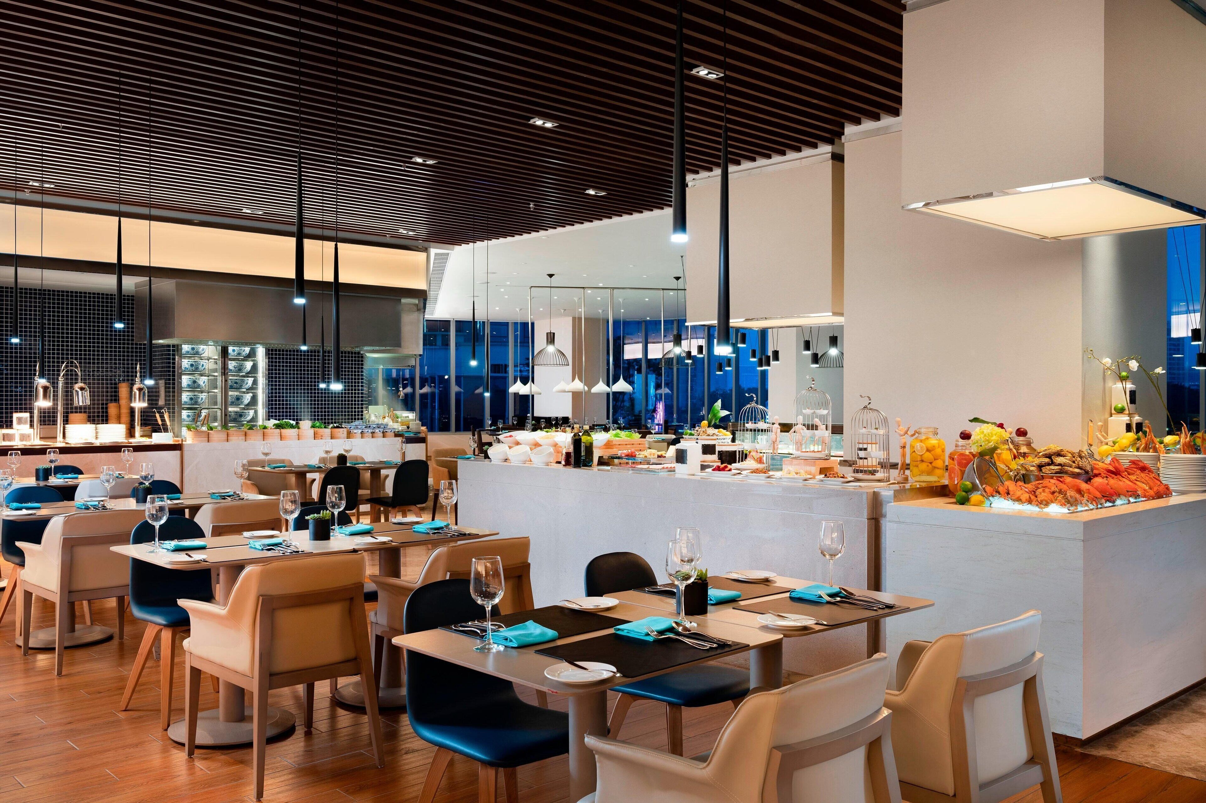 A modern restaurant with wooden flooring, blue chairs, and a central island with food and drinks.