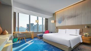 A modern hotel room with a large bed, a desk, and a view of the city skyline through large windows.