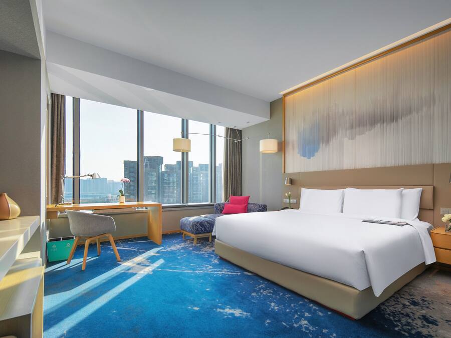 A modern hotel room with a large bed, a desk, and a view of the city skyline through large windows.