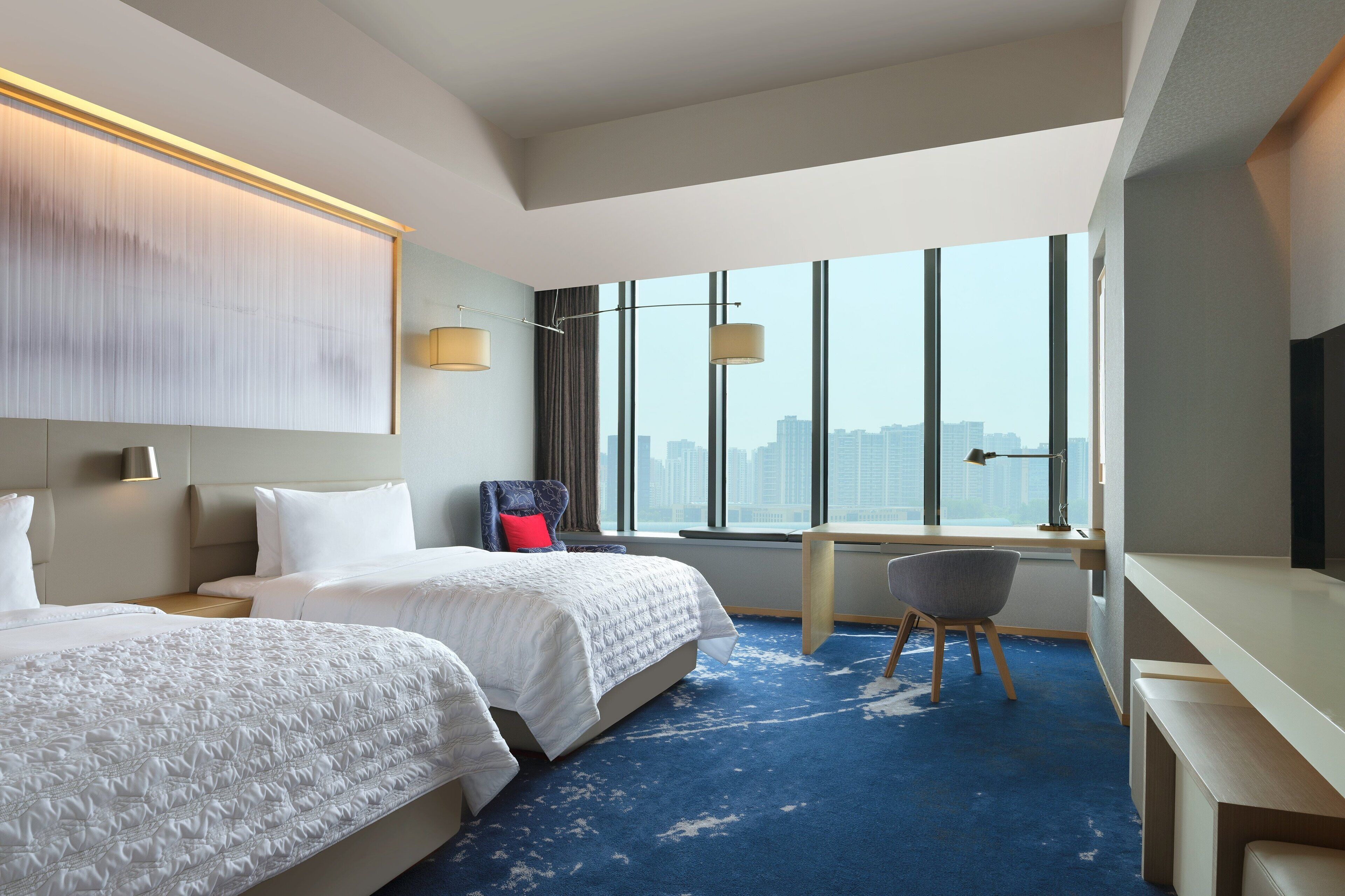 A hotel room with two beds, a desk, a chair, and a large window offering a city view.