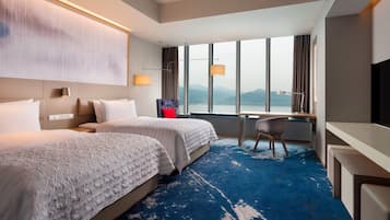 A hotel room with two beds, a desk, a chair, and a large window with a view of mountains.