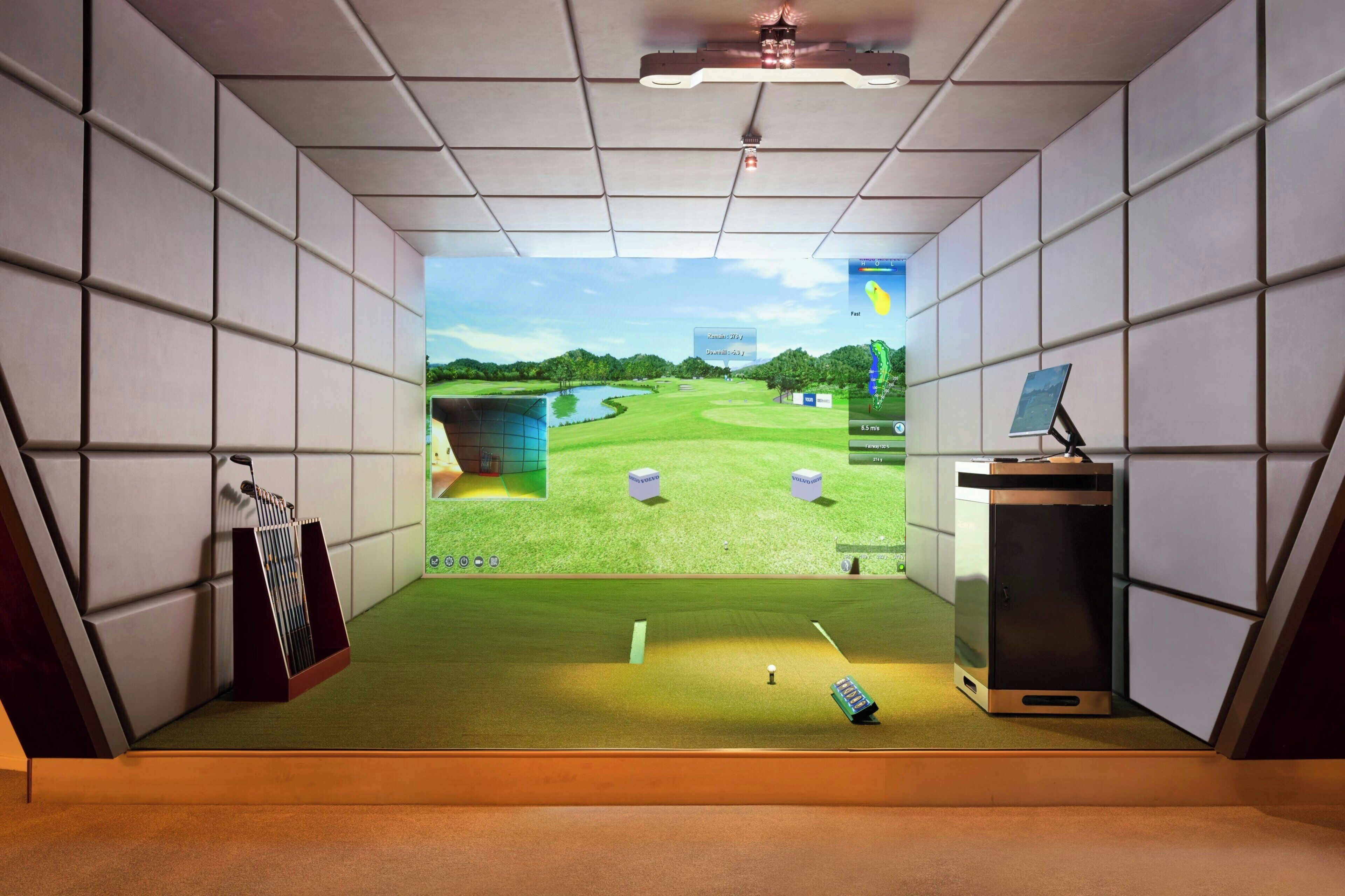 A room with a large screen displaying a golf course, a small desk with a computer, and a golf bag.