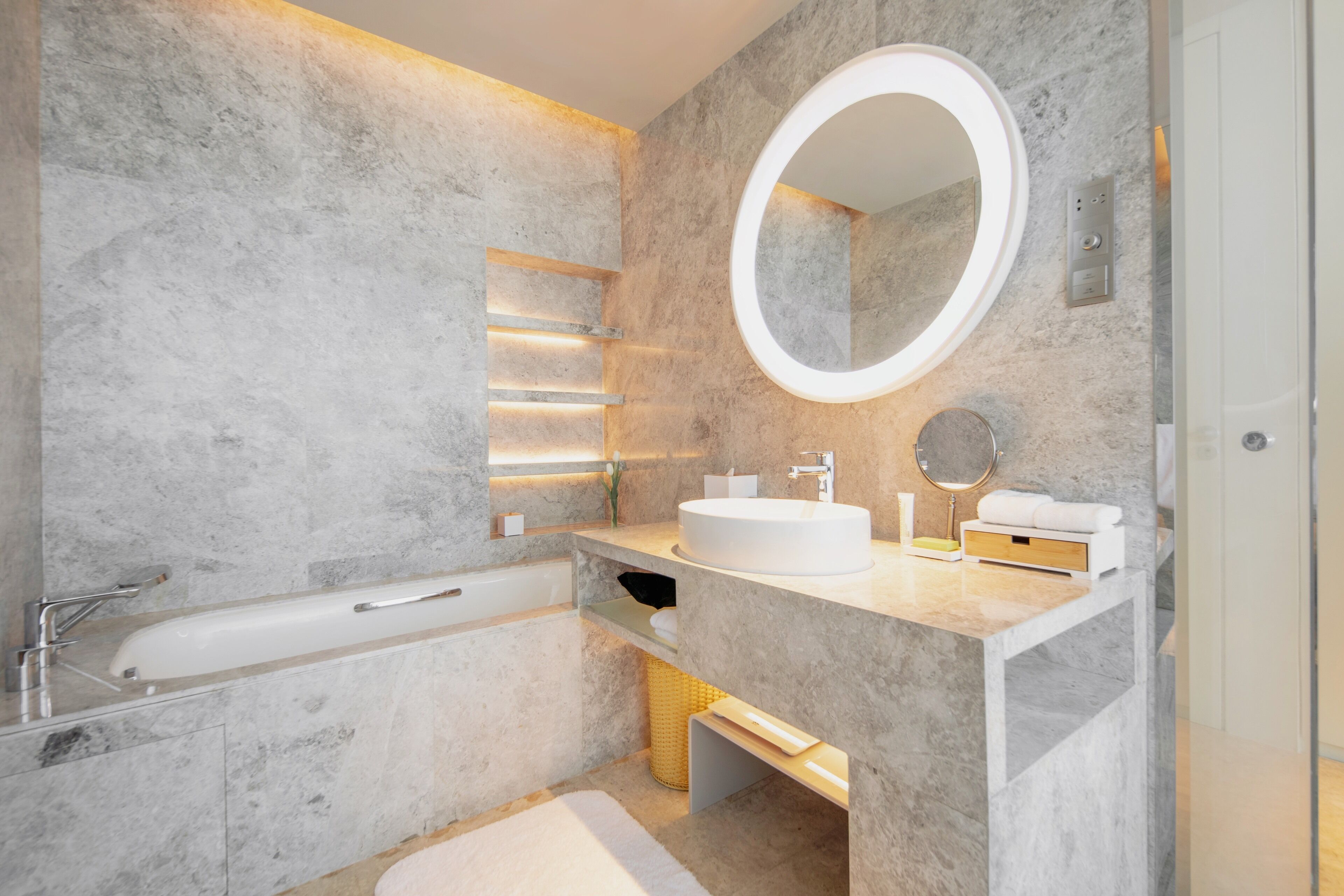 A modern bathroom with a marble wall, a freestanding bathtub, a round mirror, and a sink with a vanity.