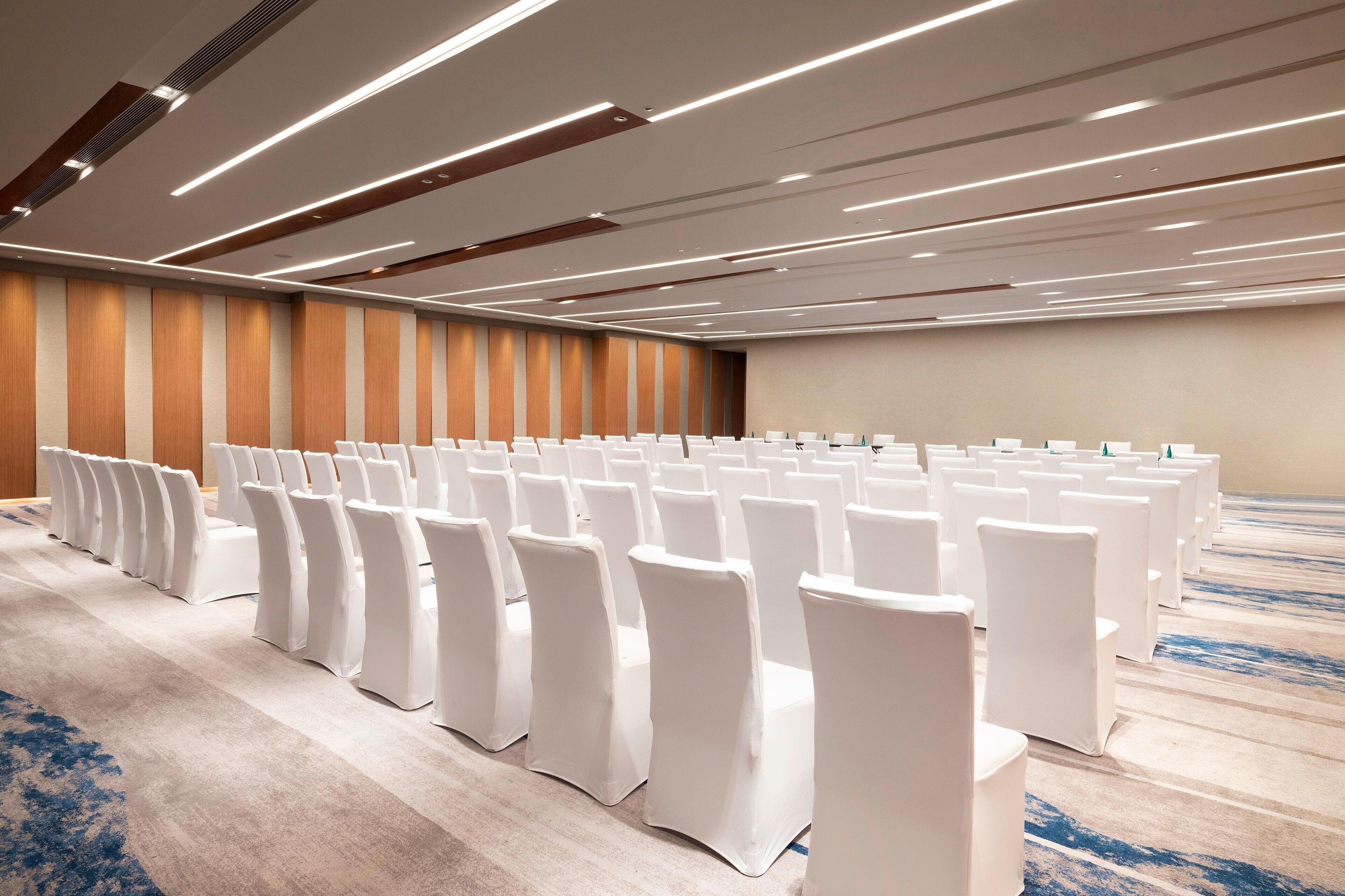 A conference room with rows of white chairs and tables, wooden paneling on the walls, and a carpeted floor.