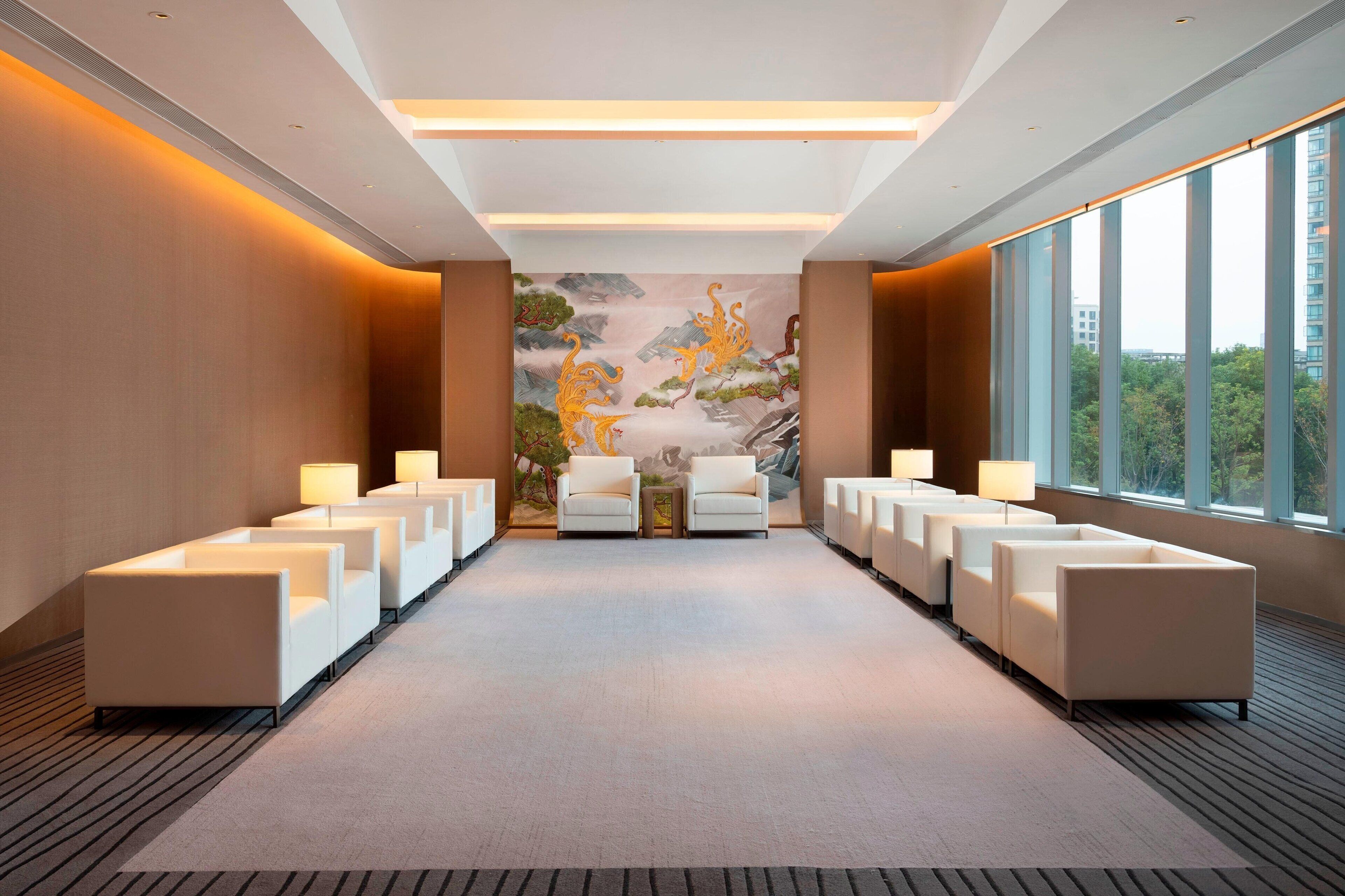 A modern hotel lobby with a long reception desk, comfortable seating, a large mural, and large windows offering a city view.