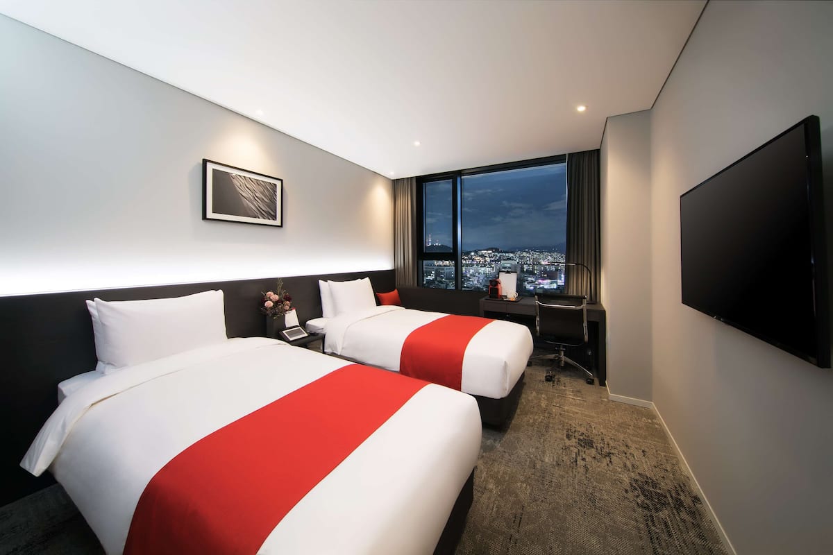 Une chambre d’hôtel avec deux lits, un bureau, une télévision et une grande fenêtre donnant sur la ville.