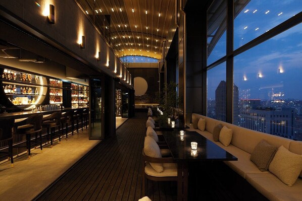 Un bar moderne offrant une vue sur la ville, doté d’un plancher en bois et d’un espace salon avec des coussins.