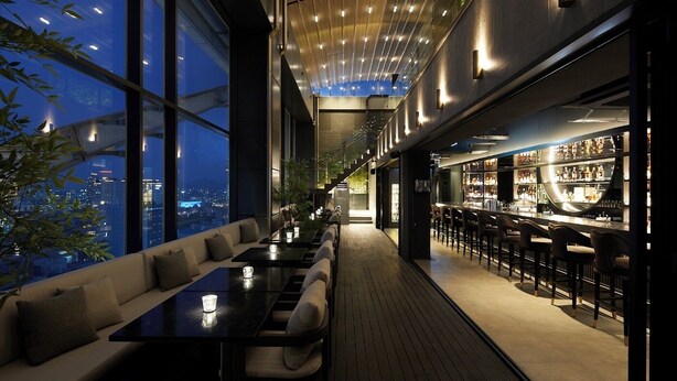 Een moderne bar met een lange toonbank en zitplaatsen links, en een bar met flessenkasten rechts.