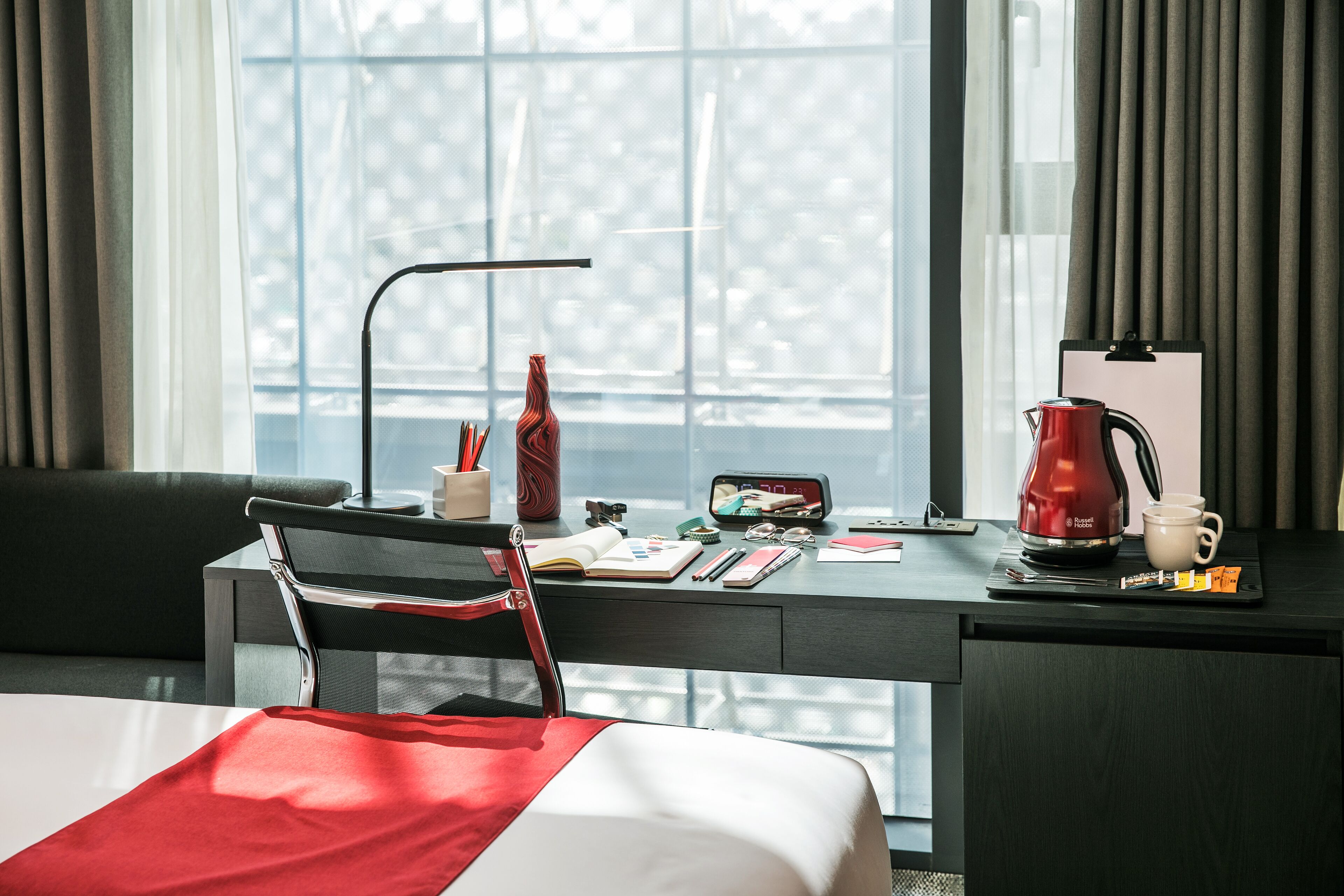 Habitación de hotel con cama, escritorio con una computadora portátil, un hervidor rojo y una ventana con cortinas.