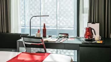 Habitación de hotel con cama, escritorio con una computadora portátil, un hervidor rojo y una ventana con cortinas.