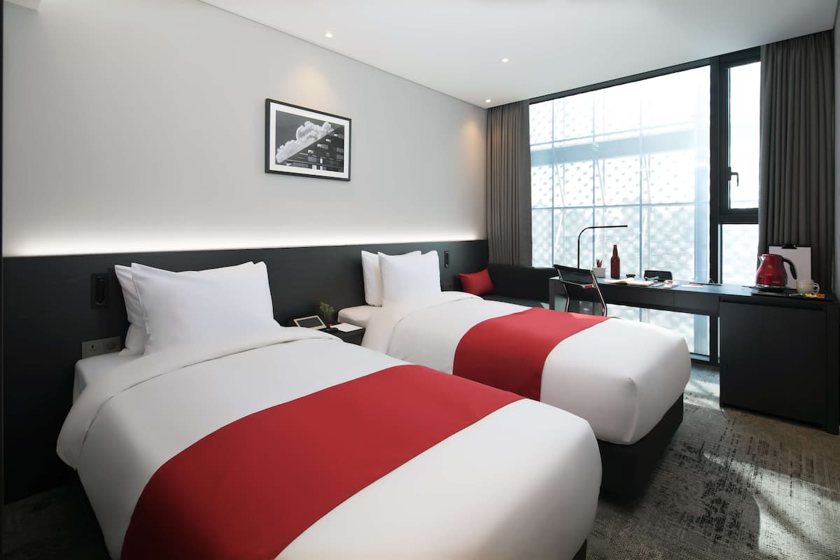 Une chambre d’hôtel avec deux lits, un bureau, une chaise, une fenêtre avec des rideaux et un tableau encadré au mur.